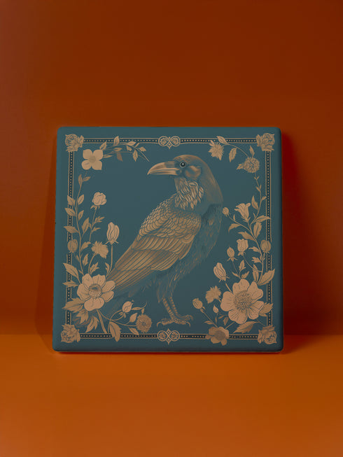 Square ceramic coaster featuring detailed gold raven illustration surrounded by botanical elements on emerald green background, with ornate border design and cork backing.